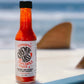 The bright red hot sauce in a bottle of Cosmic Sass Fermented Chili Sauce contrasts the baby blue ocean in the background and the baby blue surfboard it's sitting on.